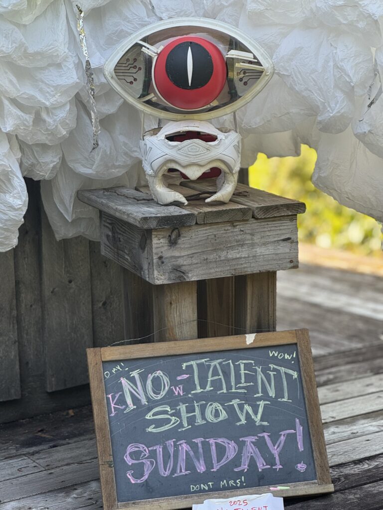 A chalkboard with an invitation to join the kNOw talent show on Sunday with a white helmet topped by an object that looks like an all seeing eye