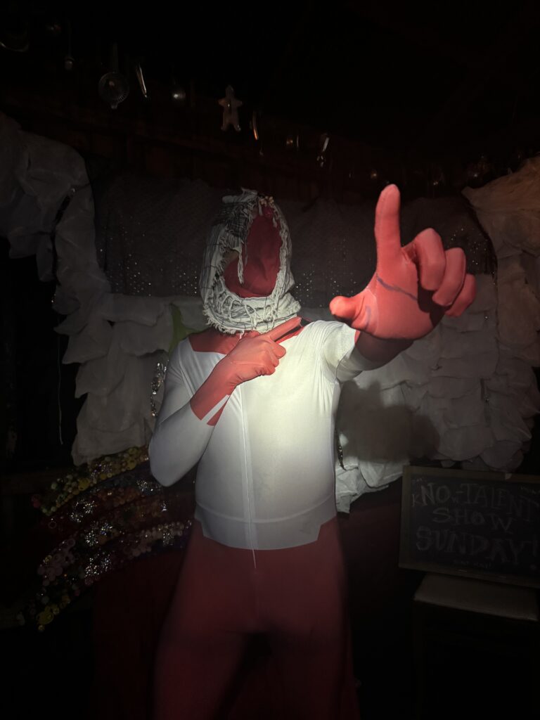 A person in a red and white unitard with a spotlight on their chest pointing up and too the right of the camera.
