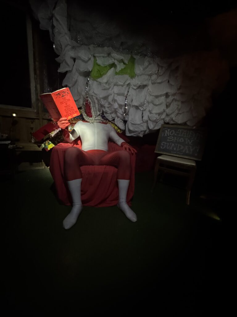 A color photo of Sully reading Faggots and their Friends between Revolutions in a red and white uni-tard with a billowy white background