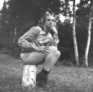 A radical faerie sitting on a birch log with one hand on their right hand on their thigh and the their chin resting on their left hand in rag drag.