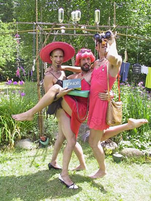 Three faeries standing together. The faerie on the left is in a pink sun hat. The middle bearded faerie is in a red slip with a pink wig and a sign in front of them with the word HONOR. The third faerie is wearing a pink dress with a green bag over their shoulder looking through binoculars.