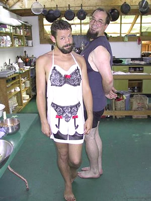 A playful photo of two men in the kitchen at the sanctuary. The man on the left has an apron on, looking down and sideways holding the apron to their front with a purple lacy and panties printed on it. The man on the right is standing just behind them to their right with their hands behind their back.
