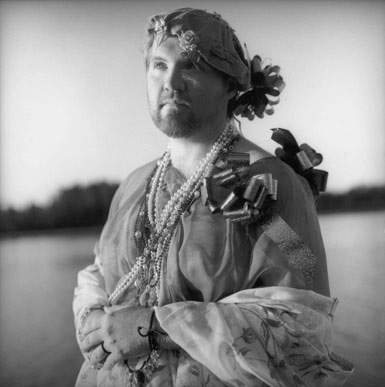A radical faerie standing in front of beaver lake with ancestor island n the background. The faerie is bearded, staring into the distance, dressed in rag drag including a big package bow on their shoulder with a shawl on wrapped around their waist and across their arms