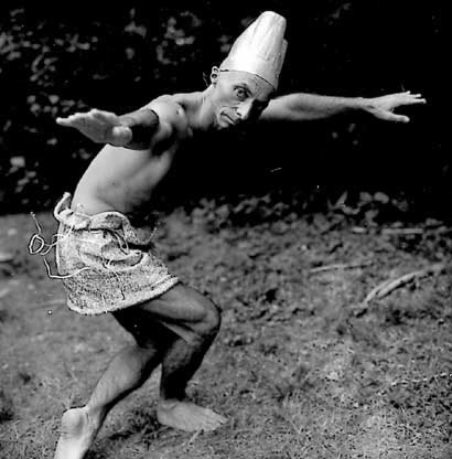 A sepia toned photo a male presenting individual, arms out stretched in curtsy with a paper fez on their head.